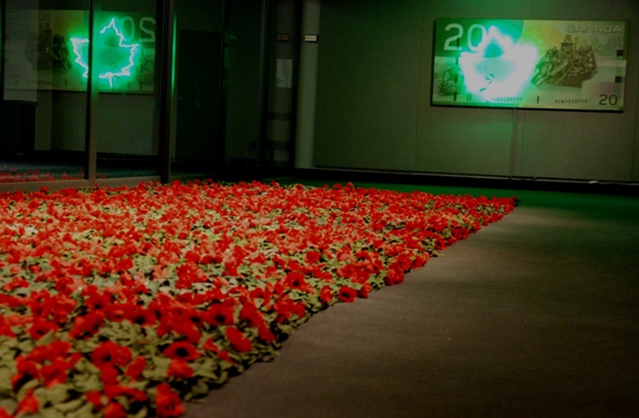 A dimly lit gallery with a large floor installation of red poppy-like flowers, green-tinted lighting, and displays showing a glowing maple leaf and a Canadian twenty dollar bill on the walls.