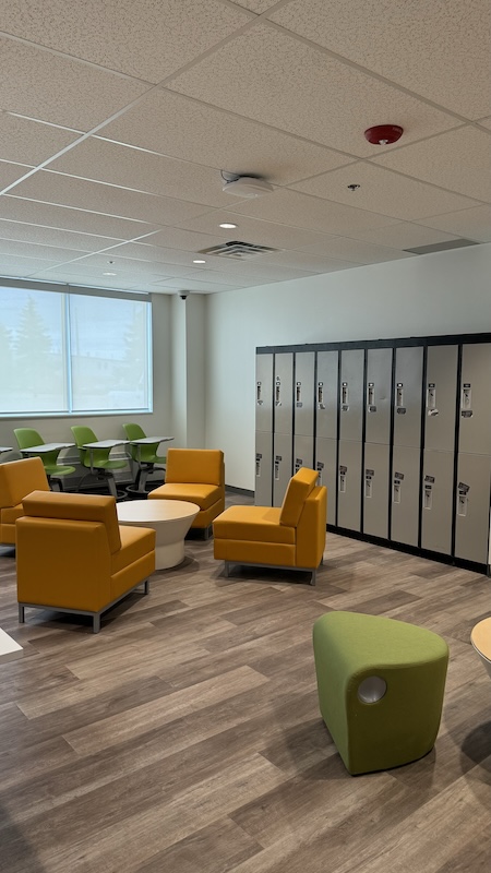 A wall of lockers behind a circle of chairs and a coffee table, as well as desks along the back wall 
