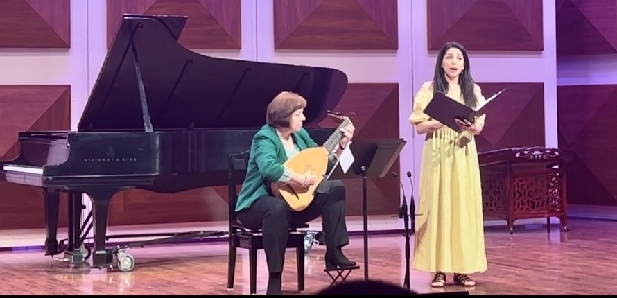 Image of young woman singing on stage, accompanied by a seated woman playing the lute