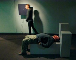 Two human figures in gallery. One on their back on a block. One walking by a wall