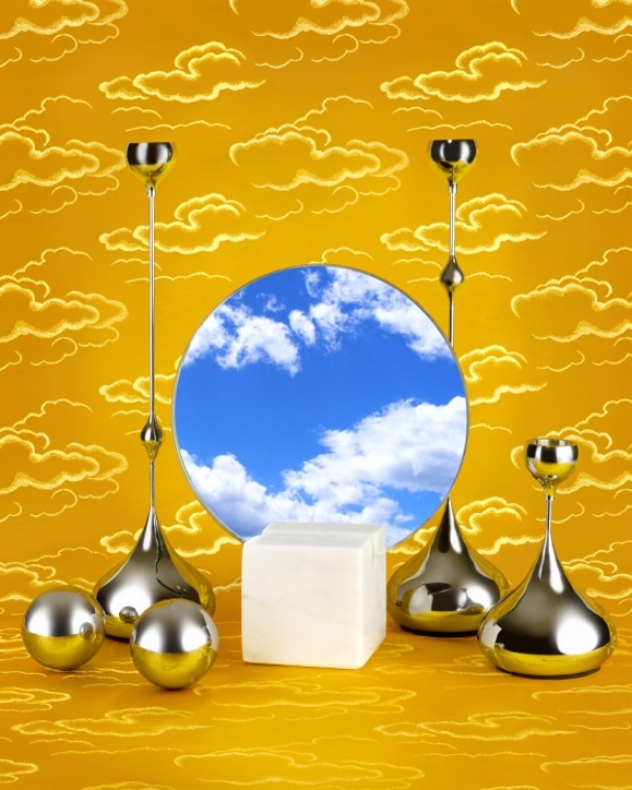 In the centre is a circular mirror with a reflection of a blue sky and cumulus clouds. To the left and right of the mirror are smooth metal spheres and abstract metal tear-shaped structures that merge into long vertical stems that become bowl-like structures. In front of the mirror and between the metal structures is a small even-textured marble cube. All the elements are on a flat, yellow cloud patterned surface and background.