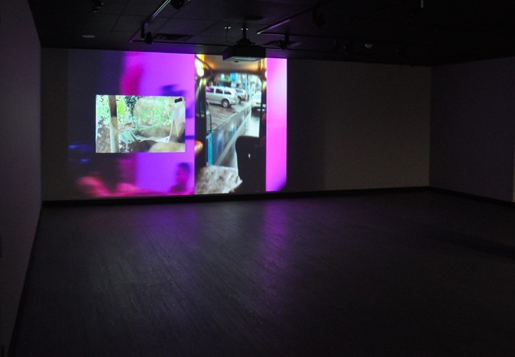 Interior of a dark gallery with a wall illuminated with overlapping images, including car on street. Unknown audio