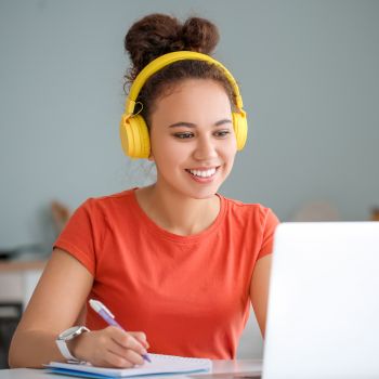 An adult learner wearing headphones, sitting in front of her laptop with a notebook, smiling and ready to learn