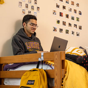 male student sitting on dorm bed typing on laptop