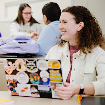 A student sitting with their laptop in a classroom. They're smiling and looking at someone out of frame. In the background there are a couple other students.