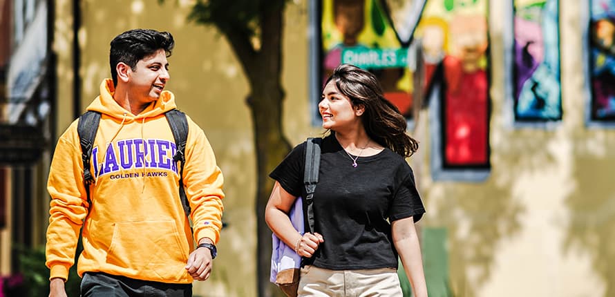 Two students walking downtown Milton. One student is wearing a gold Laurier hooded sweatshirt, the other a black short sleeve shirt. They're smiling at each other.