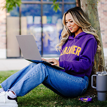female student wearing purple laurier hoodie sitting on the grass looking at laptop
