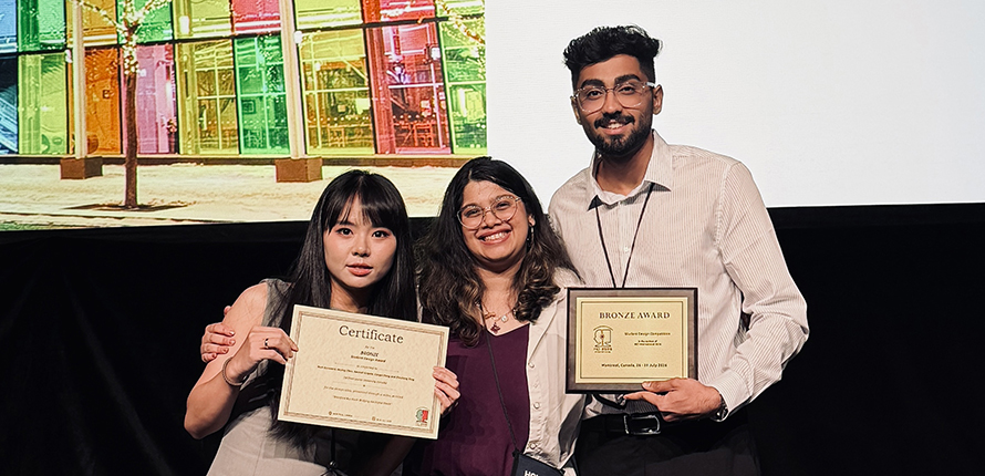 Three Master's students hold a bronze award plaque and certificate while standing on a stage